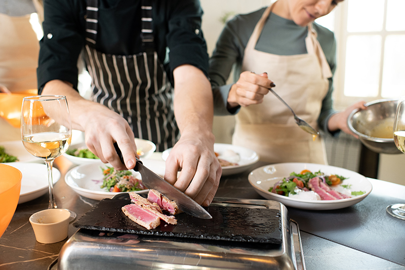 Hands of young male cooking coach chopping piece of smoked beef on special board by table among plates with served vegetable salad