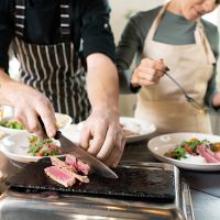 Hands of young male cooking coach chopping piece of smoked beef on special board by table among plates with served vegetable salad